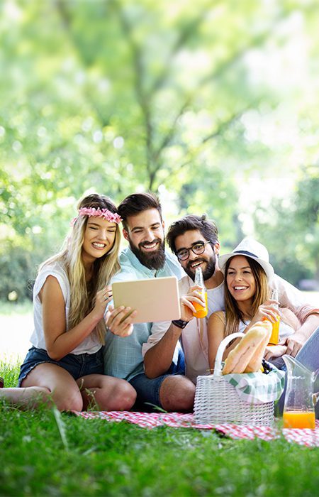 Friends in a park sitting on a blanket having a picnic looking at a tablet smiling.