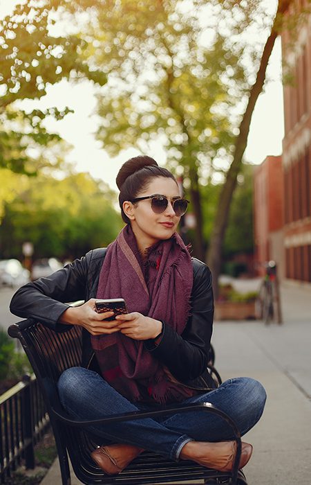 Woman sitting criss cross on a bench holding her phone wearing sunglasses, jacket and scarf.